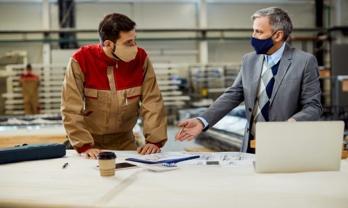 Mid adult businessman and male worker discussing about project plans while wearing protective face masks at carpentry workshop.